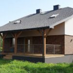 Zdjęcie przedstawia nowoczesną, piętrową elewację mieszkalnego budynku, z częściowo drewnianą werandą i balkonem.
